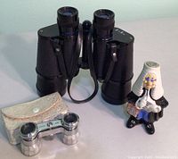 Front view of black textured binocular-flasks, small mother of pearl opera glasses with cream case, and figurine liqueur bottle with white wig and blue pants