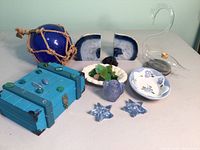 Full group shot of all items on table showing cobalt blue float glass ball, agate bookends, sea glass in white bowl, blue star-shaped glass pieces, ceramic plates, clear glass vessel, small blue pitcher, and wooden box with stone detailing.