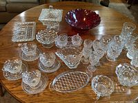 Photo showing wide assortment of clear Fostoria glassware including cups, saucers, plates, serving pieces arranged on a wooden table with large red bowl at top center.