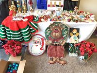 Wide view of Christmas items on table and floor including crocheted tree skirt, wall hangings, faux poinsettias, and many ornaments