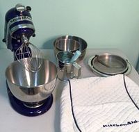 Full view of purple KitchenAid mixer with attached stainless steel mixing bowl, additional mixing bowls, cake and pie plates, hand sifter, and quilted cover on table.