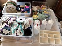 Three large plastic tubs filled with various types of ribbons on spools in multiple colors and materials, several white plastic squeeze bottles, and black plastic storage pots arranged on a table.