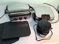 Three kitchen appliances on a counter: Toastmaster waffle maker, Griddler Gourmet panini grill, Hamilton Beach appliance for pancakes or eggs, with extra grilling plates beside them.