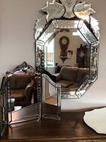 View of ornate Venetian-style mirror with floral and vine etched details along beveled edges, next to standing folding mirror with three hinged panels and wooden frame.