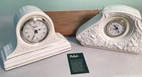 Two white porcelain Beleek mantle clocks with a small wooden wall shelf behind them.