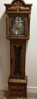 Front view of the full tall wooden grandfather clock showing bonnet top, clock face door, pendulum case with glass window, and base cabinet.