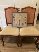 Two vintage carved wooden chairs with cane backs and beige padded seats, with a square wooden wall clock placed in front.