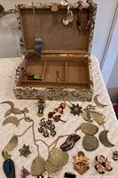 Open tapestry jewelry box showing interior compartments and various costume jewelry pieces including necklaces and pins placed in front and inside the box.