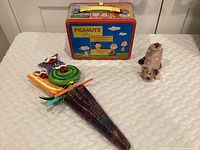Full shot showing Peanuts lunchbox, purple glass vase with three painted creatures on sticks inside, and small ceramic dog.