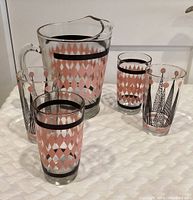 Full view of the glass pitcher along with four drinking glasses displaying pink and black diamond patterns and stylized linear tree designs.