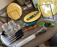 Photo showing three yellow ceramic plates on black metal holders, glass canisters, glass bowls, black-handled knives, a metal spatula, a turquoise slotted spatula, and various kitchen utensils on a round table.