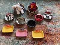 Top-down image showing a variety of pet food dishes, water bowls, and two plush toys on a carpet.