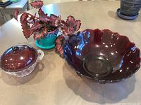 Overall photo of red glass serving bowl, candy dish with lid, and colorful red begonia plant in blue pot arranged on table