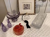 Lot displayed on a white quilted surface showing all items, including textile art print, purple candy dish, red swan dish, two small purple perfume bottles, atomizer, and cut glass dish.