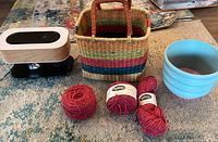 Photo showing yarn threader device, colorful woven basket, four skeins of yarn, and blue ceramic pot on carpet.