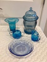 Image of six blue glass items including bright blue vase, creamer, sugar bowl, light blue covered candy dish, blue jar with lid, and low cut glass dish.