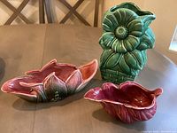 Three ceramic items on a table: pink pitcher, green vase in flower shape, flower-shaped dish with color gradient