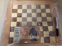 Wooden chess board with tan and dark brown squares alongside the original Milton Bradley Company Chessmen box, slightly wrapped in plastic.