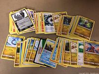 Photo showing multiple stacks of Pokémon trading cards spread on a surface including Pokémon, energy, and trainer cards, estimated around 100 cards.