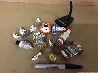 View of NASA and space-themed toys spread on beige surface, showing astronaut figurines, Apollo capsule, lunar module, space station, lunar rover, and moon rock models, with a black stand and Sharpie pen for scale.