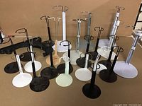 Photo showing multiple metal doll or figure display stands in black, white, and one light green, with round bases and adjustable clamps