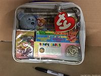 Clear vinyl collector bag with white trim and zipper containing vintage Ty Beanie Babies items including blue plush bear, sealed membership cards, newsletter, coin, and collector cards.
