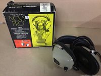 Photo showing the white ear cup headphones with black cushioning and coiled cord beside original black and yellow box with classical bust illustration.