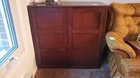 Front view of dark wood cabinet with double doors closed placed beside a window and upholstery chair.