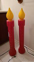 Full view of two red plastic candle decorations with yellow flame tops standing on floor next to wooden furniture and wall.