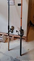 Two fishing rods standing upright with reels attached, one with cork handle and the other orange colored rod.