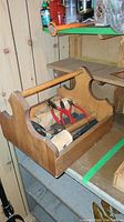 Wide view of the wooden tool bin with handle, containing various hand tools inside placed on a shelf with wood-paneled wall behind.