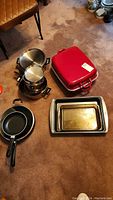Photo showing stacked metal pots and saucepans alongside a red roasting pan with lid, frying pan, and three metal baking trays.
