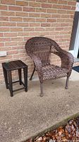 Full view of brown woven rattan armchair and square side table placed outside on concrete in front of brick wall