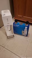 Left side view of white SodaStream carbonation machine next to box containing two carbonating bottles.