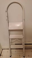 Front view of the white folding step ladder with three perforated steps and rounded upper handle.
