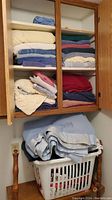 Wide view of wooden cabinet with four shelves containing folded towels in various colors: cream, white, blue, pink, red, navy, and beige. Below cabinet is a white laundry bin filled with light blue folded bedding.
