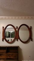 Full view of the double oval mirror mounted on the wall above a piece of furniture, showing the two connected oval mirrors with wooden framing and decorative finials.