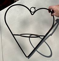 Heart shaped cast iron plant holder shown held in hand for scale on white table. Shows frame and circular pot holder ring.