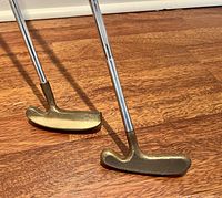Two right handed golf putters with brass mallet heads and metal shafts standing upright on wood floor.