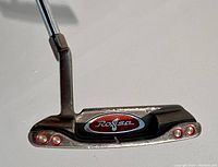 Photo showing the back of the putter head with the Rossa logo and three red circular dots on each side, highlighting the metal finish.