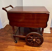 Overall side view showing the tea cart's rectangular body, wheel design with large rear wheels resembling wagon wheels and smaller castor front wheels, turned wood legs and handle.