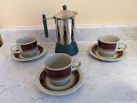 Photo showing the Italian modernist Moka espresso coffee maker with three Norwegian coffee cups and saucers, all arranged on a white countertop.