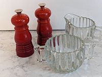 Two red Le Creuset salt & pepper grinders with Penfolds branding and two ribbed glass pieces (milk pitcher and sugar bowl) on marble surface.
