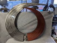 Full view of the modernist metal wall sculpture showing its entire curved form with silver and copper-tone halves, metal rods crossing the center, and polished sphere at the base.