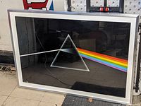 Photo showing the framed Dark Side of the Moon poster with iconic prism and rainbow light design in a silver frame, resting on a tiled floor.