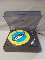 Dust cover open; yellow and blue fish-graphic vinyl placeholder, platter and S-shaped tonearm visible