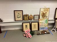 Wide shot showing 6 framed art pieces, some leaning, placed on shelf against wall, with pink faux flowers and wall-mounted metal decor