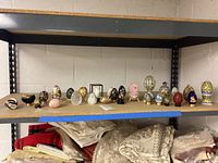 Shelf display of approximately 20 assorted decorative eggs made of various materials and sizes from 2-6 inches tall.