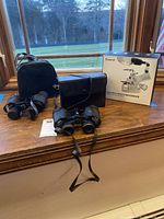 Two pairs of binoculars, their carrying cases, and a boxed Fancii USB and battery magnifier are shown on a wooden surface near window.