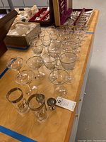 Collection of various Bohemian crystal glasses including wine glasses, cocktail glasses, and highball glasses arranged on a wooden table. Some items are stored in wooden boxes on the table.